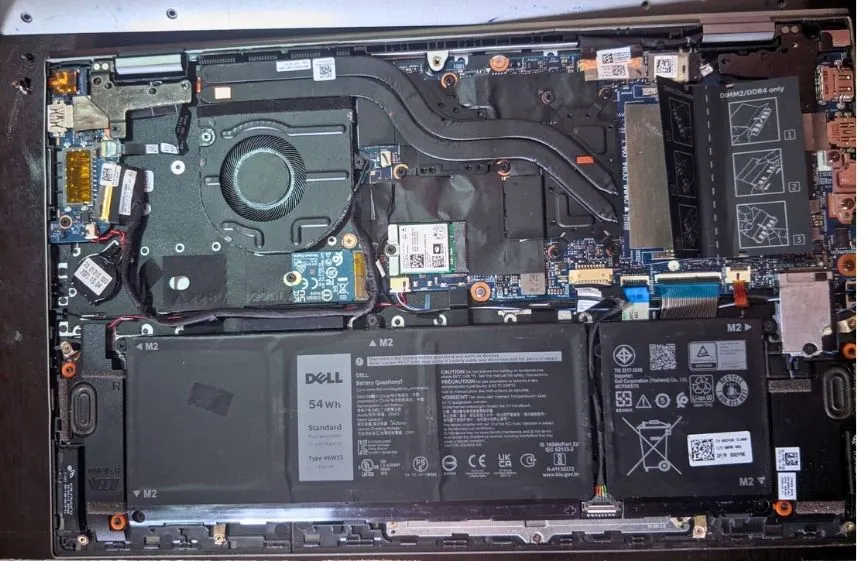 Internal view of a laptop showing the cooling fan, heat pipes, battery pack, storage slots, and motherboard components